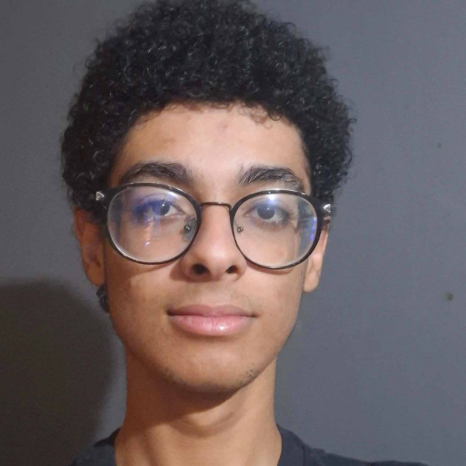 Júlio César, aluno de Design, posando em um ambiente interno com um fundo escuro. Ele usa óculos redondos e tem cabelos cacheados, com uma expressão neutra.