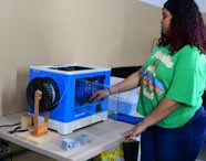 Aluna do curso de Design da Unoeste realiza atividade prática com impressora 3D azul e branca sobre uma mesa. Ela usa camiseta verde estampada e jeans, interagindo com o equipamento em ambiente de laboratório criativo, com materiais de apoio ao fundo.