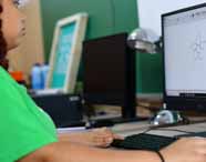 Aluna do curso de Design da Unoeste utiliza computador de mesa para desenhar uma flor digitalmente, exibida na tela. O ambiente possui luminária metálica, quadro verde ao fundo e materiais de design, demonstrando a prática de ilustração digital em sala de aula.