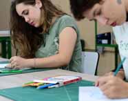 Duas alunas do curso de Design da Unoeste desenham à mão sobre folhas brancas, utilizando lápis de cor e régua. Estão sentadas lado a lado em mesa com kit de materiais artísticos, concentradas em atividade criativa durante aula prática.