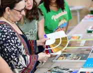Professora do curso de Design da Unoeste apresenta cartela de cores para duas alunas, analisando opções cromáticas em sala de aula. Sobre a mesa, amostras de materiais e livros de design reforçam o estudo prático das combinações visuais.