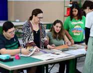 Grupo de estudantes do curso de Design recebe orientação da professora enquanto analisa catálogos de materiais, cartelas de cores e amostras sobre a mesa. O ambiente é amplo, com mesas verdes e clima colaborativo típico de aulas práticas.