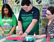 Professora acompanha dois alunos do curso de Design em atividade prática, onde utilizam estiletes e cola para criar maquetes. Ao fundo, parede decorada com pôsteres de design, ressaltando o ambiente criativo e inspirador da Unoeste.