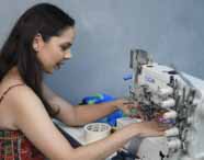 Estudante do curso de Design de Moda utiliza máquina de costura Elgin para trabalhar tecido estampado colorido. Na mesa, fita adesiva e materiais de apoio. A aluna veste vestido floral e está concentrada na atividade prática, evidenciando aprendizado técnico em ambiente acadêmico.