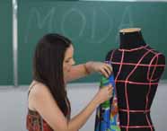 Aluna do curso de Design de Moda prende tecido azul estampado em manequim preto com marcações rosas, diante de quadro verde com a palavra "MODA" escrita. Cena retrata etapa de modelagem e criação de peças em aula prática da instituição.