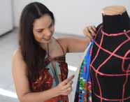Estudante mede tecido azul estampado ajustado sobre manequim preto com fitas rosas, usando fita métrica. Vestida com vestido floral, realiza atividade de modelagem em sala clara, típica das aulas práticas do curso de Design de Moda na instituição.