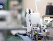 Close lateral de máquina de costura industrial branca, com detalhes metálicos e cones de linha preta ao fundo. Tecido jeans sendo costurado, foco nos mecanismos da máquina. Ambiente típico de laboratório têxtil utilizado por estudantes de Design de Moda.