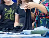 Professora segura tecido preto durante explicação prática para duas estudantes, todas em volta de mesa com materiais de costura. Professora usa vestido estampado colorido, enquanto alunas vestem camisetas pretas. Aula dinâmica do curso de Design de Moda, focada em técnicas têxteis.