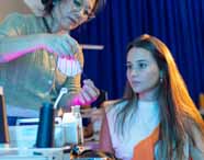 Professora orienta estudante de Design de Moda sobre uso de máscara de tecido listrado, segurando agulha e linha. Aluna observa atenta, sentada junto à máquina de costura industrial. Iluminação azul e rosa destaca ambiente moderno de ensino prático na área de moda.