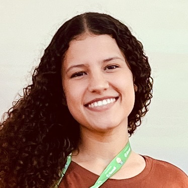 Amanda, aluna do curso de zootecnia, trajando uma blusa marrom e um crachá verde pendurado no pescoço. Ela está em um ambiente interno com fundo claro e neutro, sugerindo um espaço acadêmico ou institucional. A iluminação suave destaca os cabelos cacheados e o crachá, reforçando o contexto educacional.