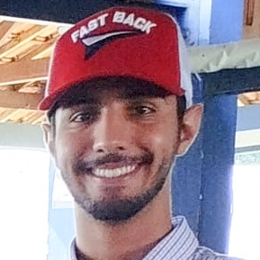 Marcelo, aluno do curso de zootecnia, trajando uma camisa listrada azul clara e um boné vermelho com a inscrição "Fast Back". Ele está em um ambiente externo coberto, com fundo formado por estruturas de madeira e metal, sugerindo um espaço rural ou técnico. A iluminação natural cria um contraste equilibrado, destacando o contexto prático.