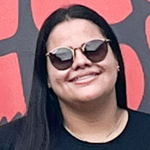 Maria Eliza, aluna do curso de zootecnia, trajando uma blusa preta. Ela está em um ambiente com fundo decorado em tons de vermelho e preto, sugerindo um espaço institucional ou de divulgação acadêmica. A iluminação uniforme destaca os cabelos lisos e o contraste entre a roupa e o fundo, reforçando o caráter educacional da imagem.