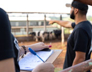 Em ambiente externo de fazenda, estudante segura prancheta com planilha e caneta, enquanto outro aponta para um grupo de bovinos ao fundo. Ambos usam camisetas escuras, sugerindo uniforme institucional. Cena representa atividade de campo do curso de Zootecnia, com foco em manejo e controle de rebanho.