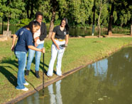 Três estudantes de Zootecnia realizam análise da qualidade da água em tanque de piscicultura. Uma utiliza sonda de medição, enquanto as outras observam e seguram equipamentos. Estão à beira do tanque, cercadas por gramado e árvores, cenário típico de práticas universitárias em aquicultura.