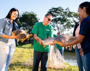 Grupo de quatro pessoas, incluindo estudantes e professor do curso de Zootecnia da Unoeste, segura grandes peixes capturados em rede à margem de tanque de piscicultura. O cenário é ao ar livre, com árvores ao fundo e clima ensolarado, ilustrando aula prática de manejo aquícola.