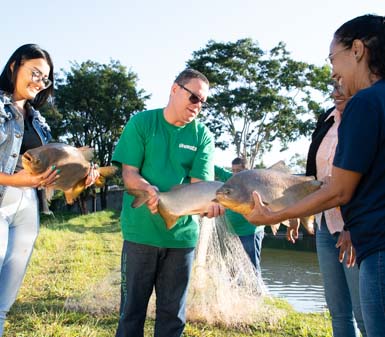 Cena externa com três pessoas, incluindo estudantes do curso de Zootecnia da Unoeste, segurando grandes peixes próximos a tanque de piscicultura. Uma mulher veste jaqueta jeans, outra camiseta azul, e um homem usa camiseta verde com logotipo da instituição. Ao fundo, árvores e céu claro completam o ambiente.
