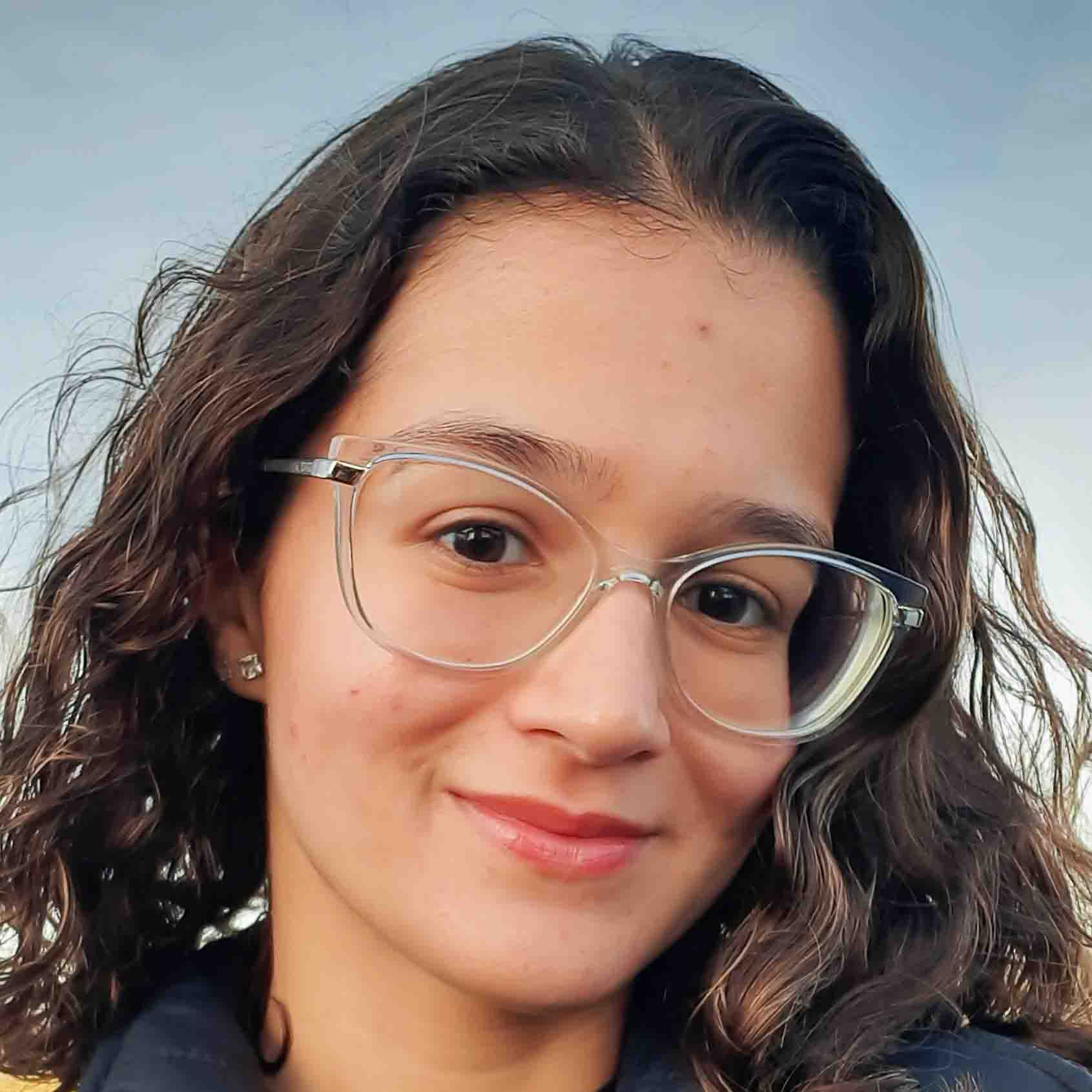 Maria Julia, aluna de Engenharia Mecatrônica, posando em um ambiente externo com um céu claro ao fundo. Ela usa óculos de armação transparente e tem cabelos cacheados, com um sorriso suave.