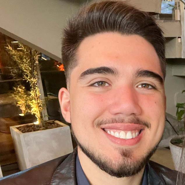 Rafael, aluno de Engenharia Mecatrônica, posando em um ambiente interno com uma escada e plantas ao fundo. Ele tem barba rala e um sorriso largo.