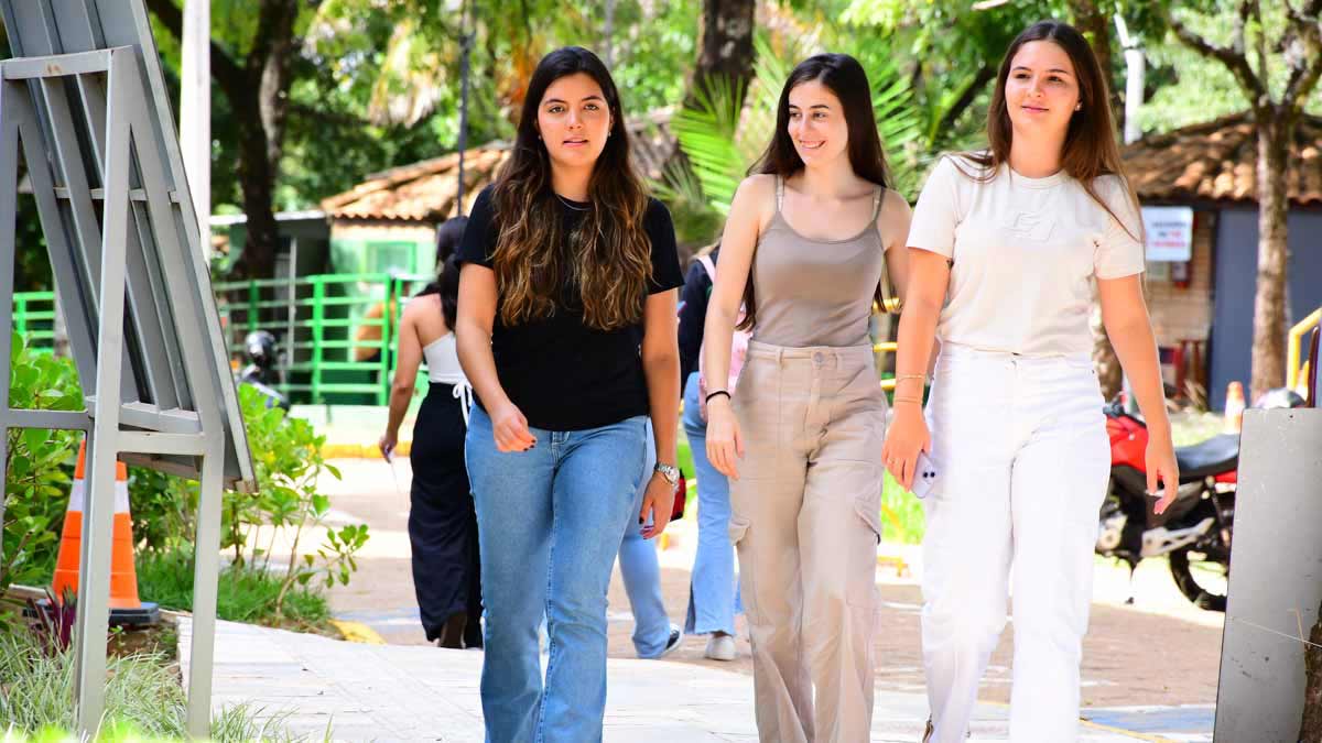Três estudantes caminham por uma alameda arborizada do campus da Unoeste, rodeadas por árvores e vegetação. As jovens usam roupas casuais em tons claros e escuros, demonstrando clima descontraído de convivência universitária no curso de Engenharia Mecatrônica.