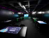 Sala de aula moderna da Unoeste, utilizada por Engenharia Mecatrônica, com mesas retangulares equipadas com telas sensíveis ao toque, cadeiras giratórias e iluminação indireta em tons roxos. Ao fundo, grande tela digital exibe interface interativa, ideal para metodologias ativas.