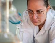 Aluna de Engenharia Química da Unoeste, usando jaleco branco, realiza experimento em laboratório. Ela manipula vidrarias científicas transparentes, como balão volumétrico e condensador, sobre bancada, demonstrando atenção aos procedimentos químicos em ambiente acadêmico.