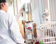 Estudante de Engenharia Química da Unoeste, com jaleco branco e óculos de proteção, opera equipamento cromado ao lado de bancada repleta de tubos de ensaio e frascos. A luz natural entra pelas persianas, destacando o ambiente moderno do laboratório de análises químicas.