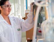 Aluna de Engenharia Química da Unoeste, vestindo jaleco branco com logotipo da universidade, manuseia equipamento metálico de laboratório. Na bancada, erlenmeyer com líquido vermelho e diversas vidrarias, evidenciando a prática experimental no curso.