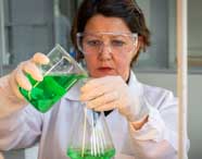 Estudante de Engenharia Química da Unoeste, usando jaleco, luvas e óculos de proteção, despeja solução verde de um béquer para um erlenmeyer. O ambiente é claro, com bancadas e vidrarias, ilustrando análise de produtos químicos em laboratório universitário.