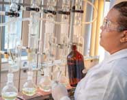 Aluna de Engenharia Química da Unoeste prepara experimento em laboratório equipado com sistema de destilação. Ela segura frasco âmbar e funil, cercada por tubos de vidro conectados e balões volumétricos, em ambiente iluminado por luz natural através das janelas.