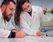 Dois estudantes de Engenharia Química realizam experimento em laboratório, ambos de jaleco branco. Um anota dados enquanto a outra ajusta instrumento de medição vertical com régua milimetrada, mostrando integração entre teoria e prática em instrumentação química.