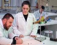 Laboratório de Engenharia Química da Unoeste com alunos em atividade prática. Em destaque, aparelho experimental montado com trilho, carrinho verde e sensores eletrônicos; estudante anota resultados enquanto outro observa, ressaltando o ensino aplicado do curso.