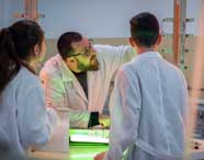 Professor de Engenharia Química da Unoeste orienta dois estudantes em laboratório, todos de jaleco branco. Eles observam equipamento de medição vertical iluminado por luz verde, interagindo com instrumentos e discutindo conceitos práticos de química aplicada.