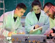 Três estudantes de Engenharia Química da Unoeste desenvolvem experimento em laboratório, utilizando jalecos brancos. Manipulam aparelhos eletrônicos, fios e vidrarias, anotando resultados. Ao fundo, quadro verde com fórmulas químicas, reforçando o ambiente acadêmico.