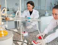 Duas alunas de Engenharia Química da Unoeste trabalham em bancada de granito, manipulando equipamentos de destilação com líquidos amarelos em balões de fundo redondo. Ambas usam jaleco e luvas, em ambiente laboratorial amplo e bem iluminado.
