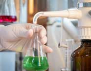 Mão enluvada de estudante de Engenharia Química segura erlenmeyer com líquido verde sob bico dosador branco acoplado a frasco âmbar. Ao fundo, outros frascos e líquidos coloridos, compondo cenário típico de laboratório de química na Unoeste.