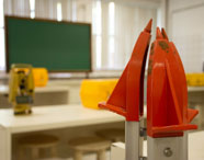 Sala de aula do curso de Geoprocessamento com foco em dois prismas topográficos laranja, usados para medições. Ao fundo, quadro verde, teodolito amarelo e caixas de transporte amarela sobre mesas brancas, compondo ambiente técnico acadêmico.