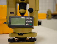 Close em teodolito digital amarelo com visor e botões, posicionado sobre mesa branca. Ao fundo, caixas de transporte amarelas e ambiente de laboratório do curso de Geoprocessamento, evidenciando estrutura moderna para aulas práticas.
