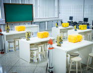 Laboratório do curso de Geoprocessamento equipado com mesas brancas, bancos, teodolitos digitais amarelos, caixas de transporte amarelas e quadros verdes. Ao fundo, computadores, formando um espaço completo para atividades práticas e teóricas.