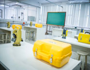 Visão ampla do laboratório de Geoprocessamento da Unoeste, com várias mesas brancas, bancos, teodolitos digitais amarelos, caixas de transporte amarelas e quadro verde ao fundo. O ambiente é iluminado e moderno, ideal para formação técnica.