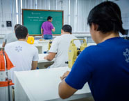 Estudantes do curso de Geoprocessamento sentados em mesas brancas equipadas com teodolitos digitais amarelos e prismas laranja, assistem a aula enquanto professor escreve no quadro verde. Ambiente típico de laboratório prático da Unoeste.