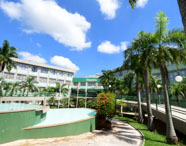 Vista externa do campus da Unoeste em Presidente Prudente, com edifícios de vários andares, jardins bem cuidados, palmeiras altas, arbustos floridos, bandeiras e fonte vazia em primeiro plano sob céu azul com nuvens.
