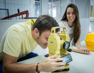 Dois estudantes do curso de Geoprocessamento da Unoeste manipulam teodolito digital amarelo sobre mesa branca, com caixa de transporte amarela ao lado. O ambiente é de laboratório, reforçando o caráter prático da formação.