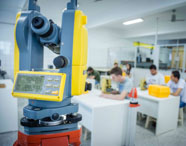 Laboratório do curso de Geoprocessamento com teodolito digital amarelo e prisma laranja em destaque à frente. Ao fundo, estudantes sentados em mesas brancas realizam atividades práticas, demonstrando aprendizado técnico na Unoeste.