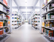 Corredor central da biblioteca da Unoeste, com estantes metálicas repletas de livros organizados por cor e categoria. Ambiente amplo, bem iluminado, próprio para estudos dos alunos do curso de Geoprocessamento e demais áreas