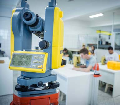 Imagem em sala de aula do curso de Geoprocessamento, com destaque para um teodolito digital amarelo e azul em primeiro plano. Ao fundo, estudantes trabalham em mesas brancas com equipamentos topográficos e cones laranja, evidenciando prática técnica na formação profissional, possivelmente na Unoeste de Presidente Prudente.