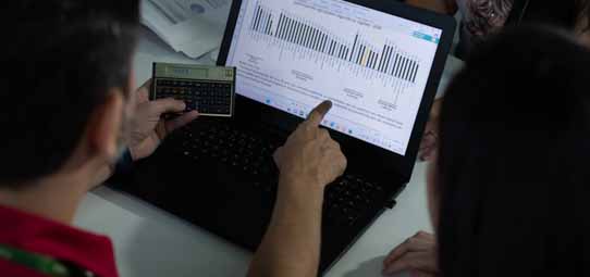 Dois estudantes analisam gráficos de barras exibidos em um notebook preto. Um deles aponta para a tela com o dedo indicador enquanto o outro segura uma calculadora científica dourada. Ao fundo, papéis sobre a mesa sugerem ambiente de estudo ou análise de dados empresariais.