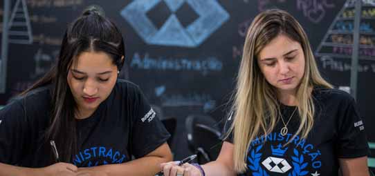 Duas estudantes de Administração sentadas lado a lado em sala de aula, concentradas fazendo anotações. Ambas vestem camisetas pretas com a inscrição "Administração" em azul e logotipo institucional. A estudante à esquerda tem cabelo liso preto e a da direita cabelo liso loiro. Ao fundo, um quadro negro decorado com desenhos de pirâmide, losangos geométricos e a palavra "Administração" em giz branco compõe o cenário da sala temática.