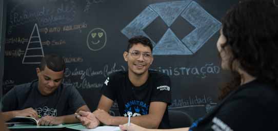 Três estudantes jovens sentados em mesa de sala de aula interagem durante atividade em grupo. Ao centro, um jovem de óculos, camiseta preta com estampa de "Administração" sorri amplamente. À esquerda, outro estudante de camiseta escura olha para baixo com caderno aberto. À direita, uma estudante de costas participa da conversa. Ao fundo, um quadro negro decorado com desenhos de pirâmide, emoji de coração, losangos e a palavra "nistração" (Administração), criando um ambiente descontraído e colaborativo.
