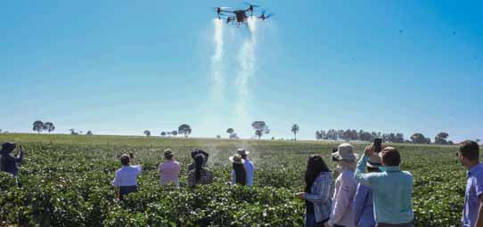 Drone agrícola de grande porte voa sobre uma extensa lavoura verde, liberando jatos de água ou defensivo agrícola pelos quatro braços da aeronave. No solo, um grupo de aproximadamente dez pessoas observa a operação, algumas fotografando com celulares. Céu azul claro ao fundo com árvores no horizonte.