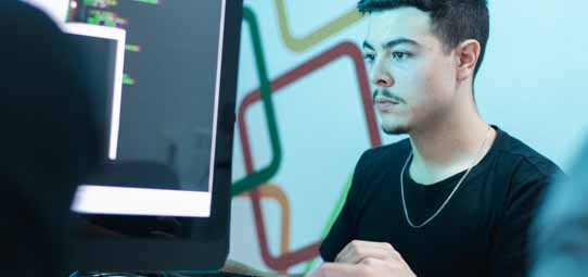 Jovem do sexo masculino, de cabelos escuros, camiseta preta e corrente fina no pescoço, olha fixamente para um monitor de computador que exibe linhas de código coloridas. Ao fundo desfocado, uma parede com formas geométricas nas cores verde, vermelho e amarelo.
